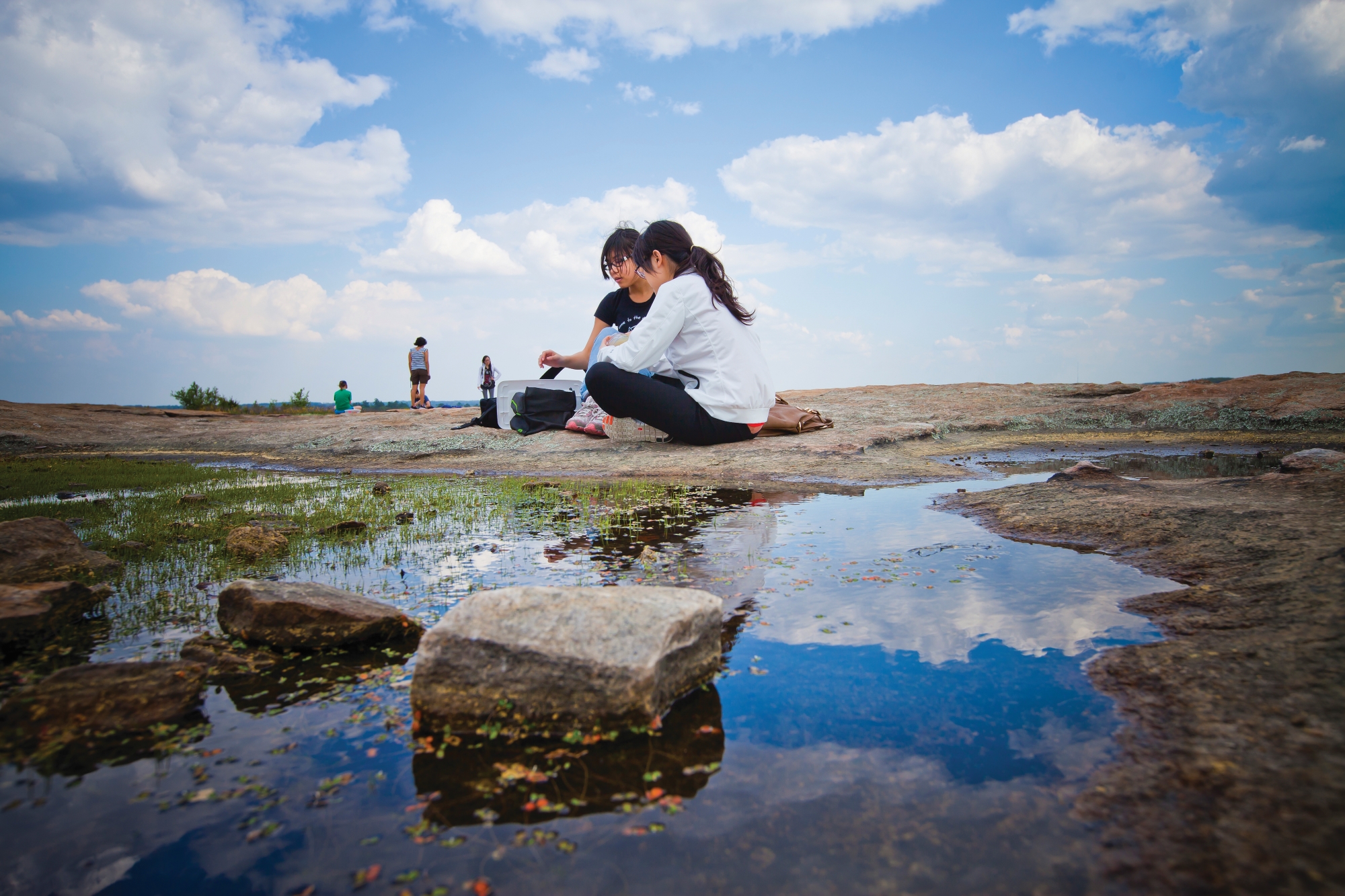 Two students sit on a mountain next to a puddle that is reflecting the image of the sky.